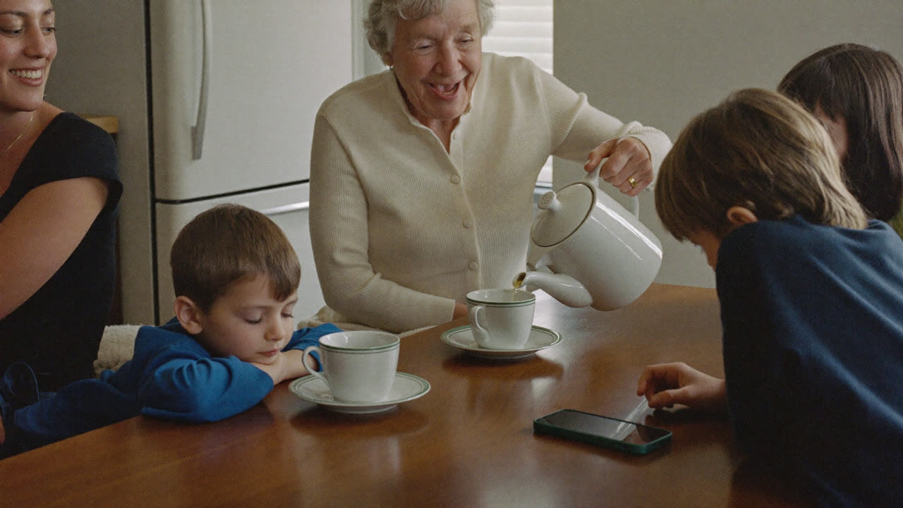 Three generations around a kitchen table, tea being poured, a phone lying flat on the table between them.